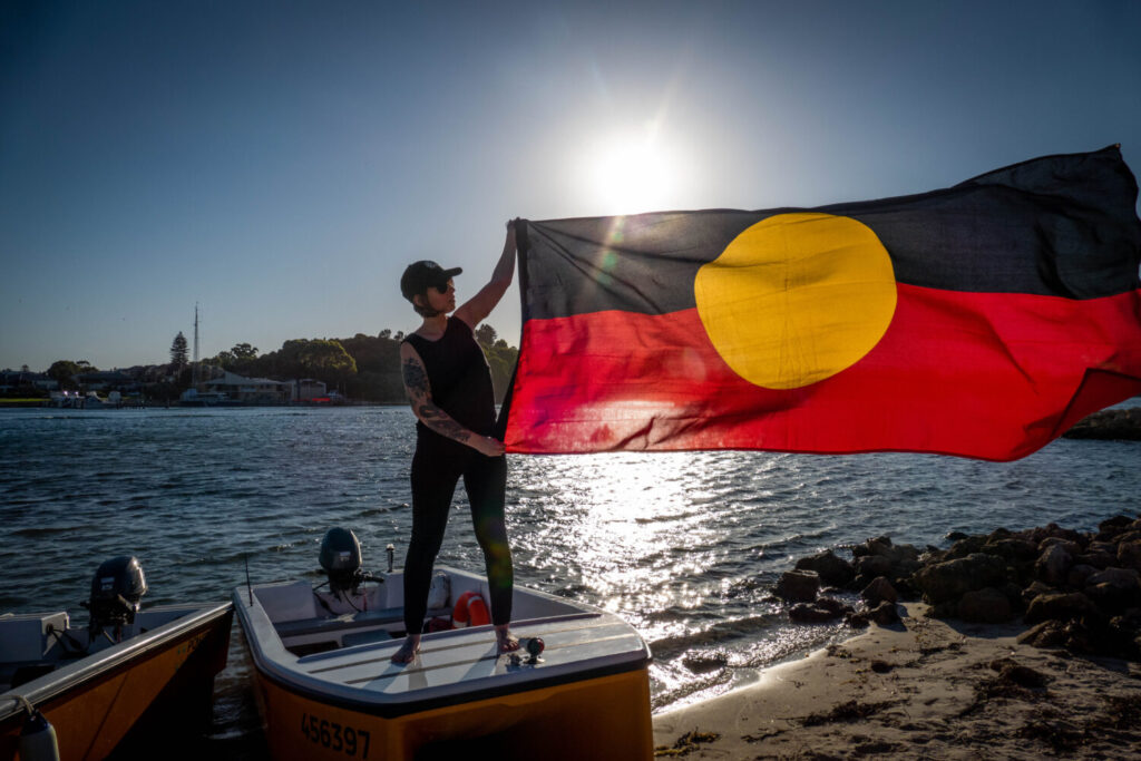 Person standing on beached boat holding the Australian Aboriginal flag